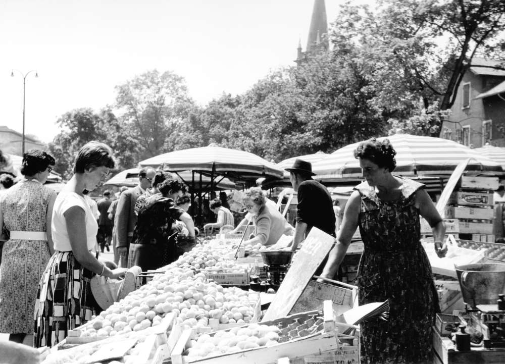 Marktstand Anfang der 1960er Jahre mit Marktfrau und Marktbesuchern. Schwarz-Weiß-Aufnahme.