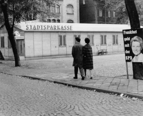 Der Höchster Marktplatz mit der ehemaligen Stadtsparkasse, die 100-jähriges Jubiläum gefeiert hat. Im Vordergrund ein spazierendes Paar sowie ein SPD-Wahlplakat von Anfang der Sechziger Jahre.